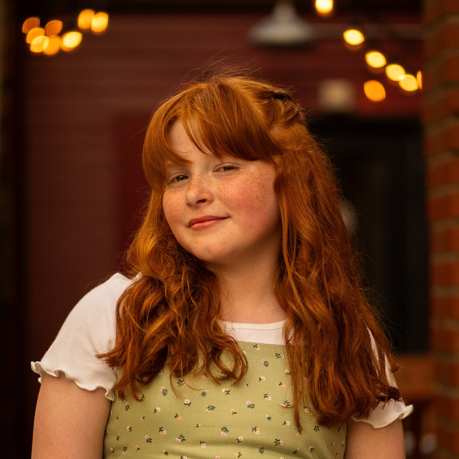 Young girl with red hair wearing a green dress with a white collar against a blurred indoor background.